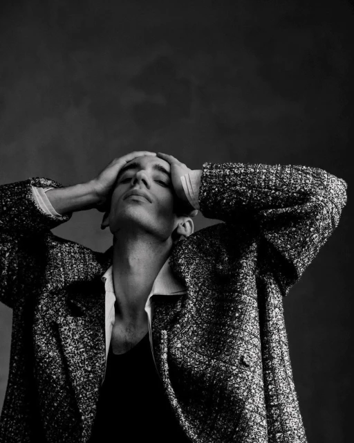 Black and white portrait of a young adult man with hands behind his head, wearing a textured coat, against a neutral backdrop.