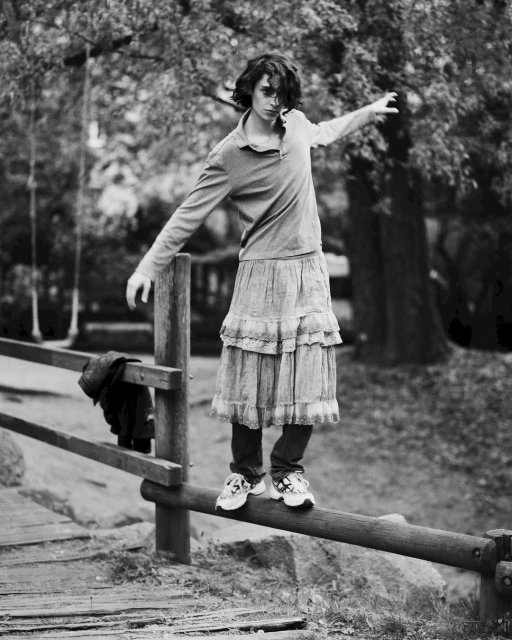 Monochrome portrait of a young adult balancing on a wooden railing in a park, wearing a dress over jeans with greenery in the background.
