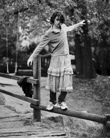 Monochrome portrait of a young adult balancing on a wooden railing in a park, wearing a dress over jeans with greenery in the background.