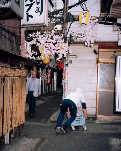 Street scene in Japan features a man pulling a cart and another man walking by buildings and cherry blossom decorations on a city street.
