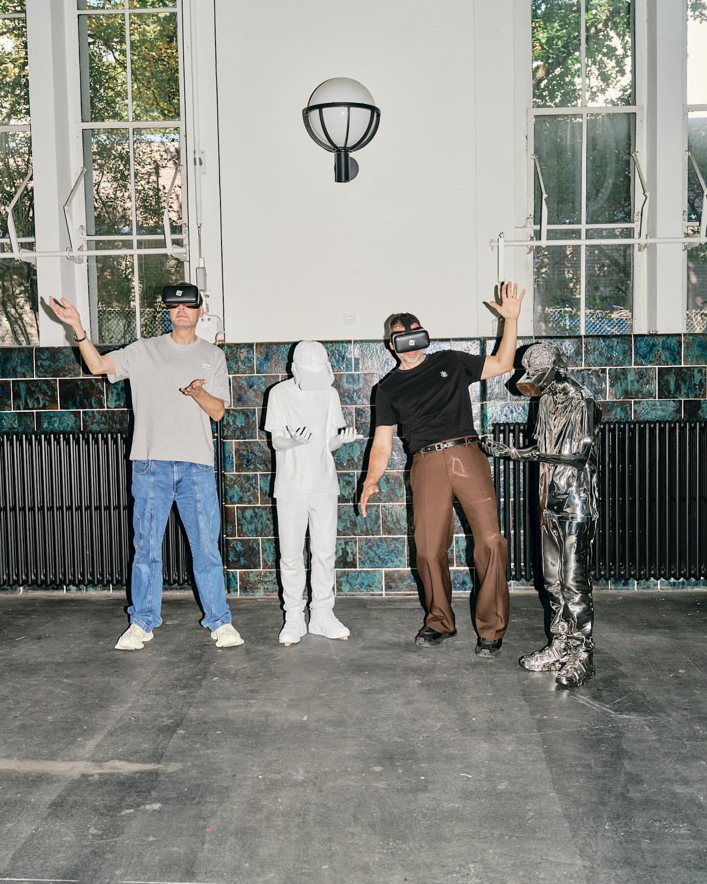 Four people, two wearing VR headsets, and two in costume posing indoors with tiled walls and window light.
