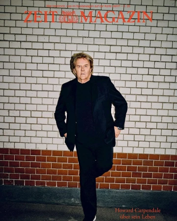 Man wearing a black suit and shirt leaning against a tiled brick wall under red magazine typography