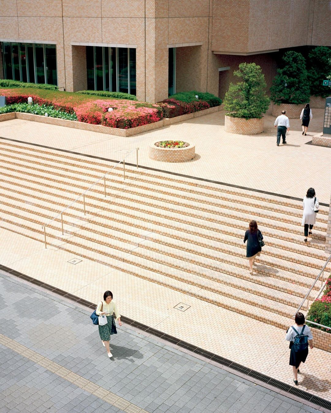 High-angle shot of several people walking on a geometrically patterned, tiered outdoor staircase in an urban environment during the daytime.
