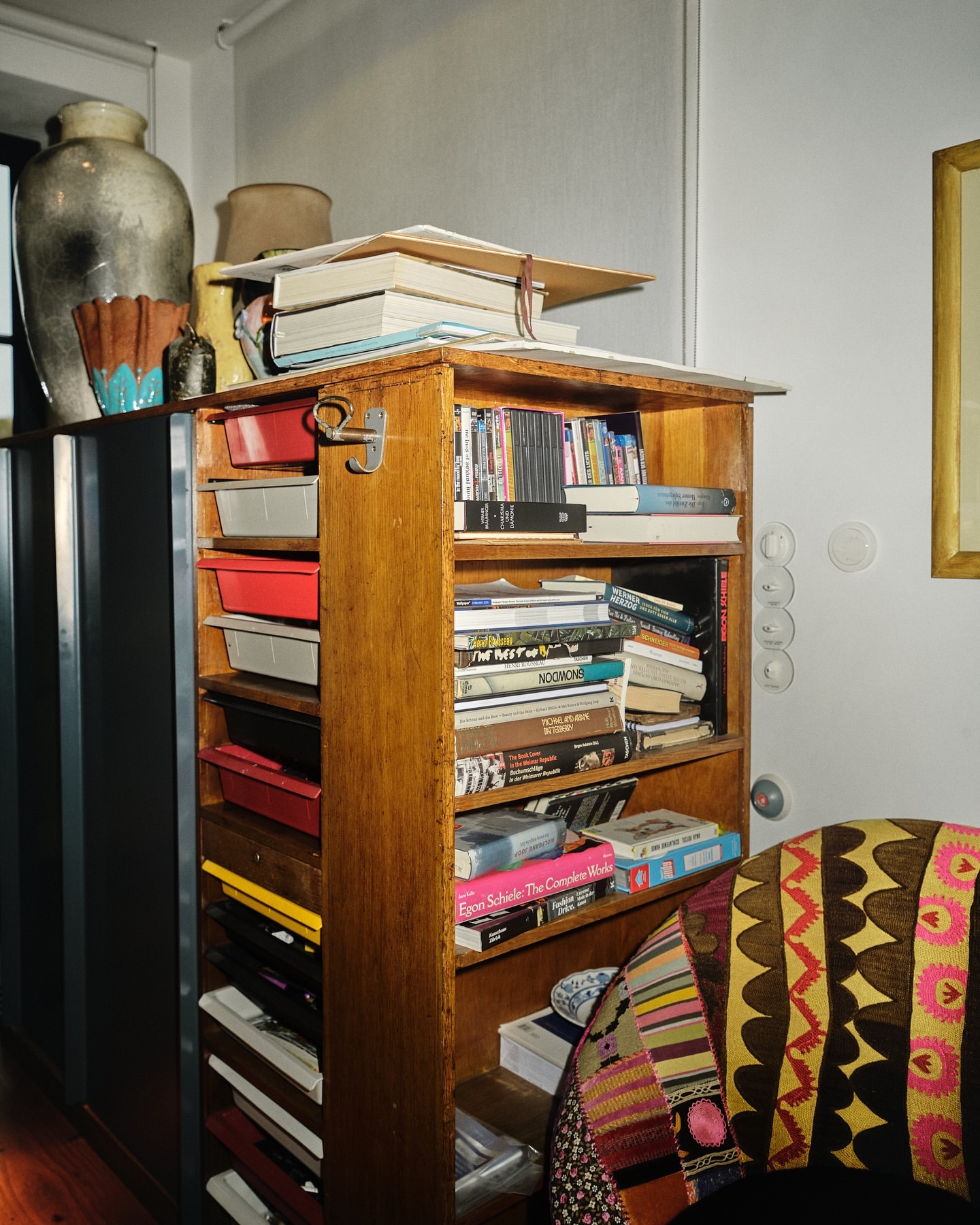 Wooden bookshelf filled with books and miscellaneous objects, against a white wall with a glimpse of a patterned chair.