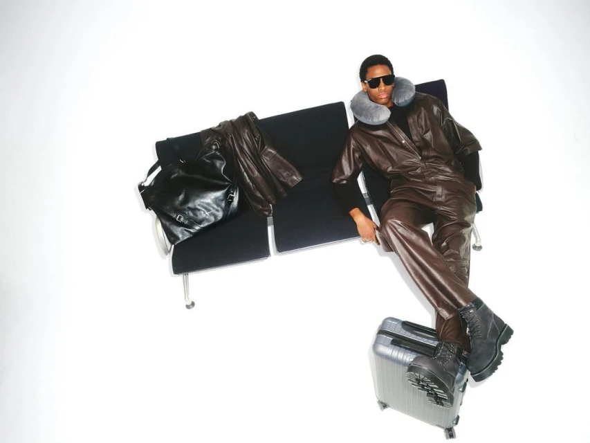 A person wearing a brown leather jumpsuit and sunglasses sits on a bench next to a black bag and silver suitcase in a white space.