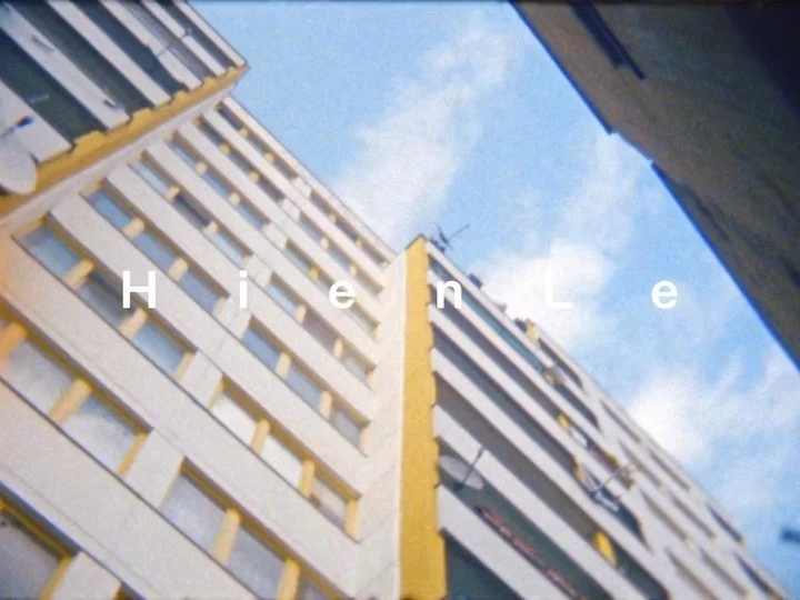 High-angle view of a tall apartment building reaching towards a blue sky with wispy clouds, partially framed by adjacent structures.
