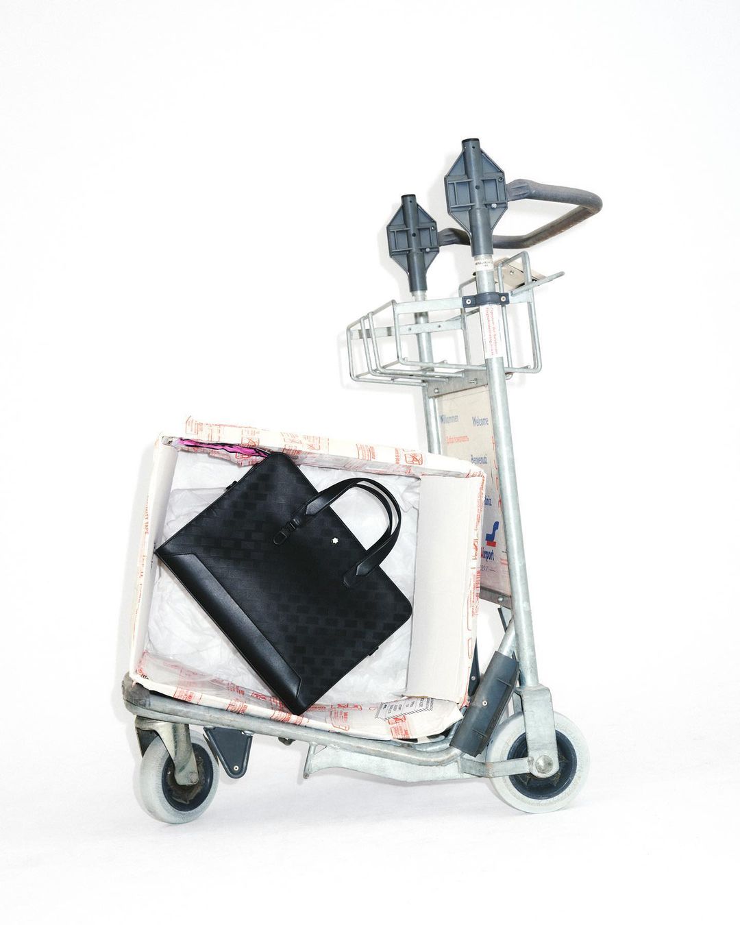 Black briefcase on a metal luggage cart filled with packing materials and covered in text patterns, set against a white background.