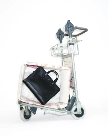Black briefcase on a metal luggage cart filled with packing materials and covered in text patterns, set against a white background.