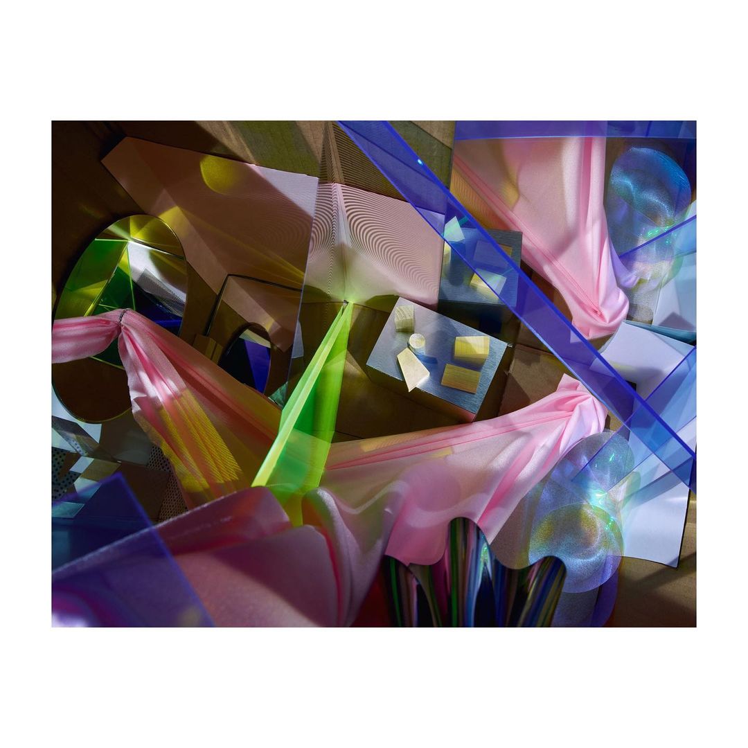Abstract still life features draped pink fabric, geometric shapes, and iridescent elements in a collage aesthetic with blue and green accents.