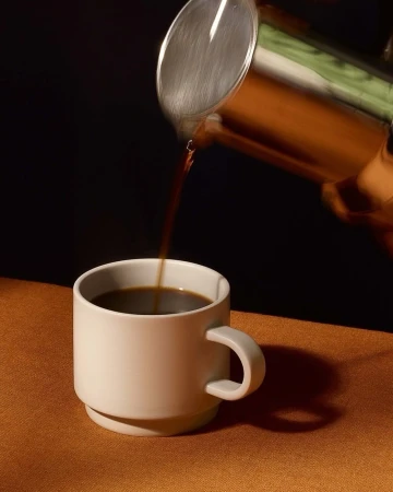 A steaming stream of dark coffee pours from a stainless steel carafe into a white ceramic mug, set against a black background.
