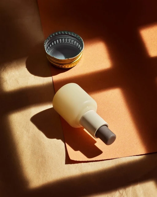 Still life of a lipstick tube and a metal cap casting window shadows on a multi-toned surface, shot with indoor lighting.