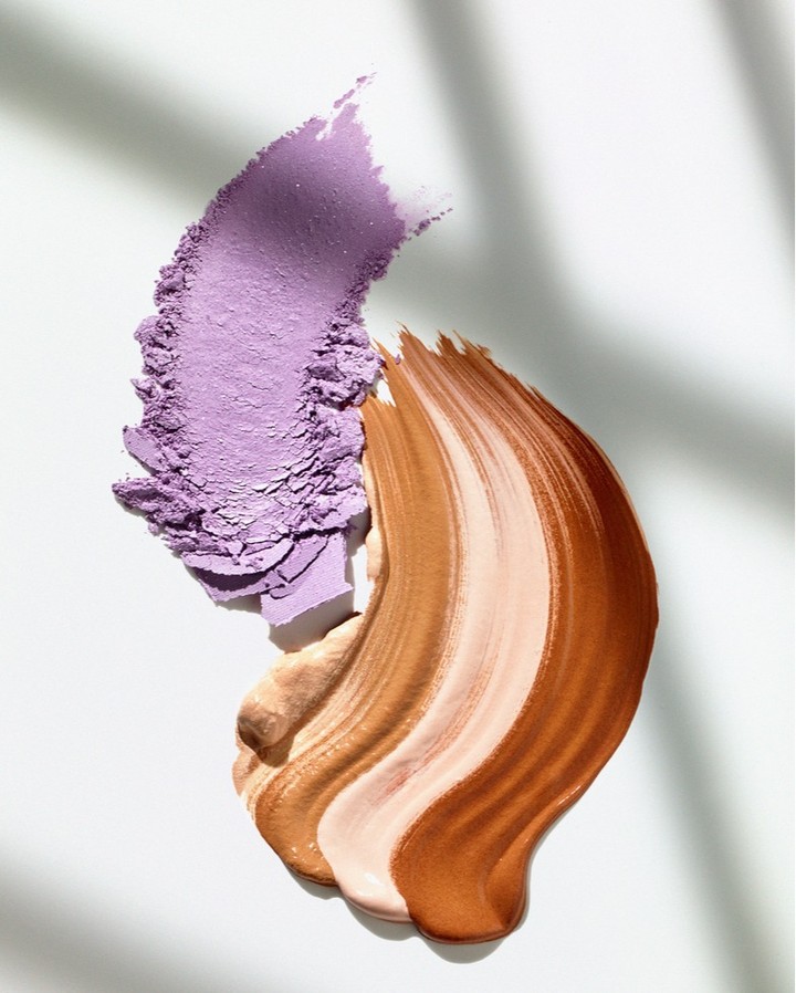 Overhead shot of beige and brown liquid foundation swatches with lilac powder eyeshadow on a white surface with window-blind shadows.