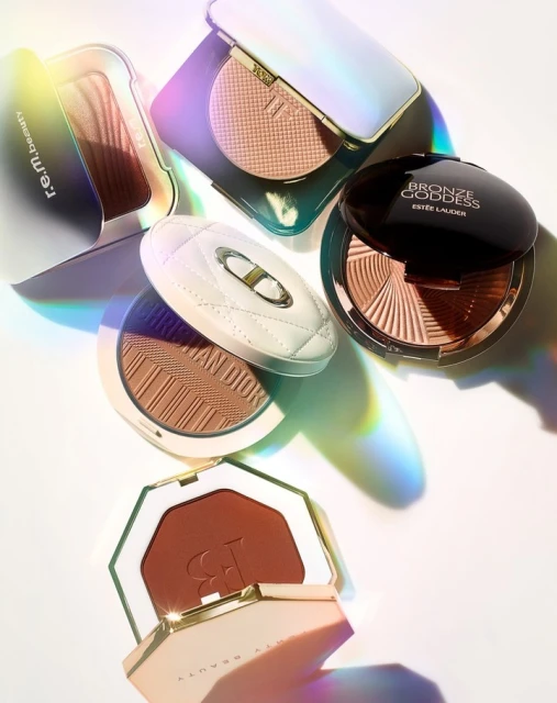 Cosmetic bronzer collection including r.e.m. Beauty, Estee Lauder, Dior, and Fenty Beauty with light reflecting off the product cases.