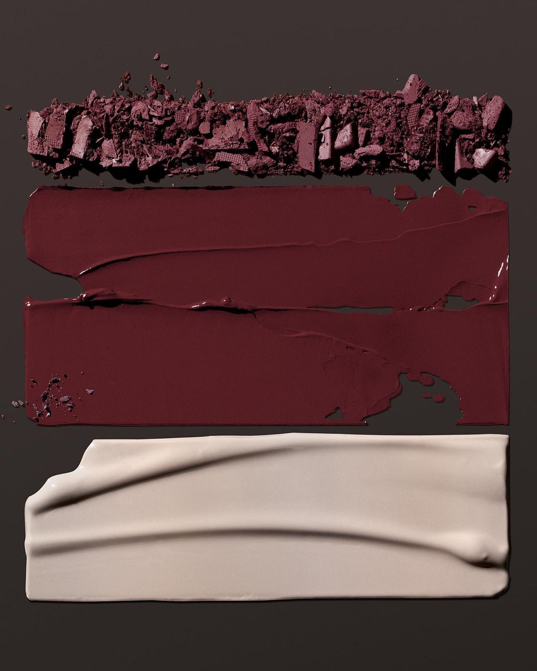 Cosmetic swatches of eyeshadow, lipstick, and foundation in a horizontal arrangement on a dark background showcasing beauty product textures.