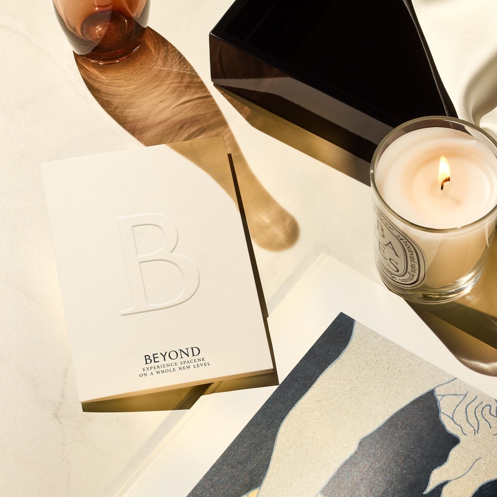 Still life photo of a candle, glass vase, and stationary with embossed text reading 'Beyond Experience Space' in warm, diffused light.