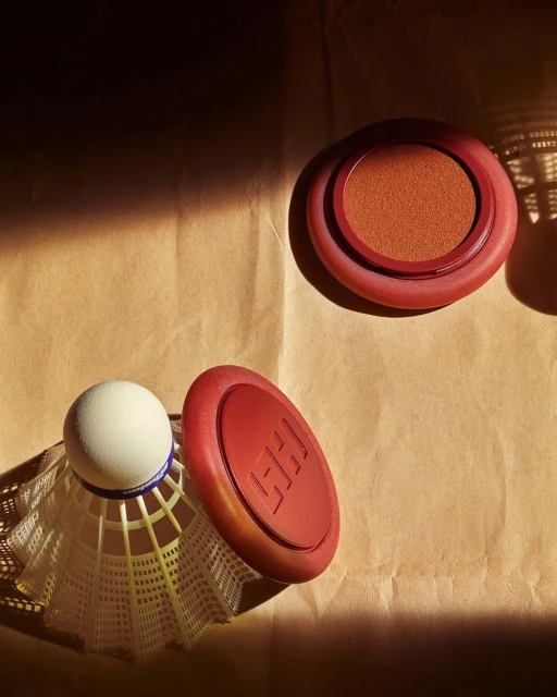 Still life featuring makeup compacts near a badminton shuttlecock on craft paper, casting soft shadows, ideal for beauty product imagery.