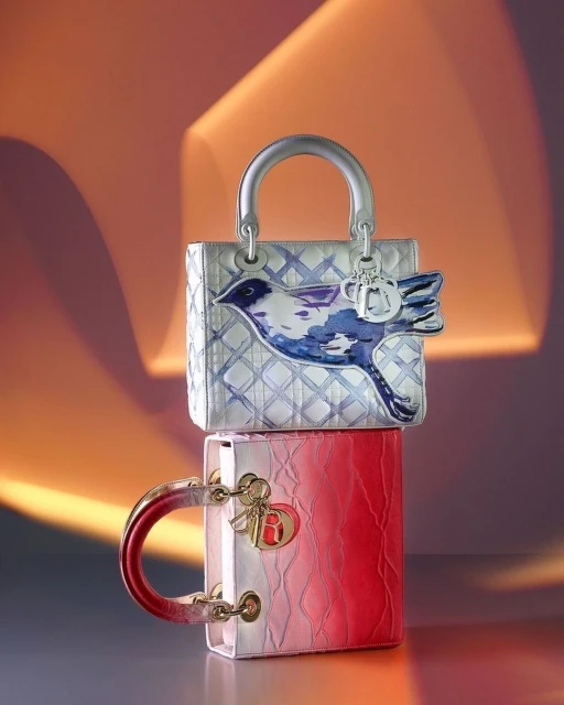 Two Lady Dior handbags stacked, the top bag is a silver and blue with bird art, and the bottom bag is pink and red.
