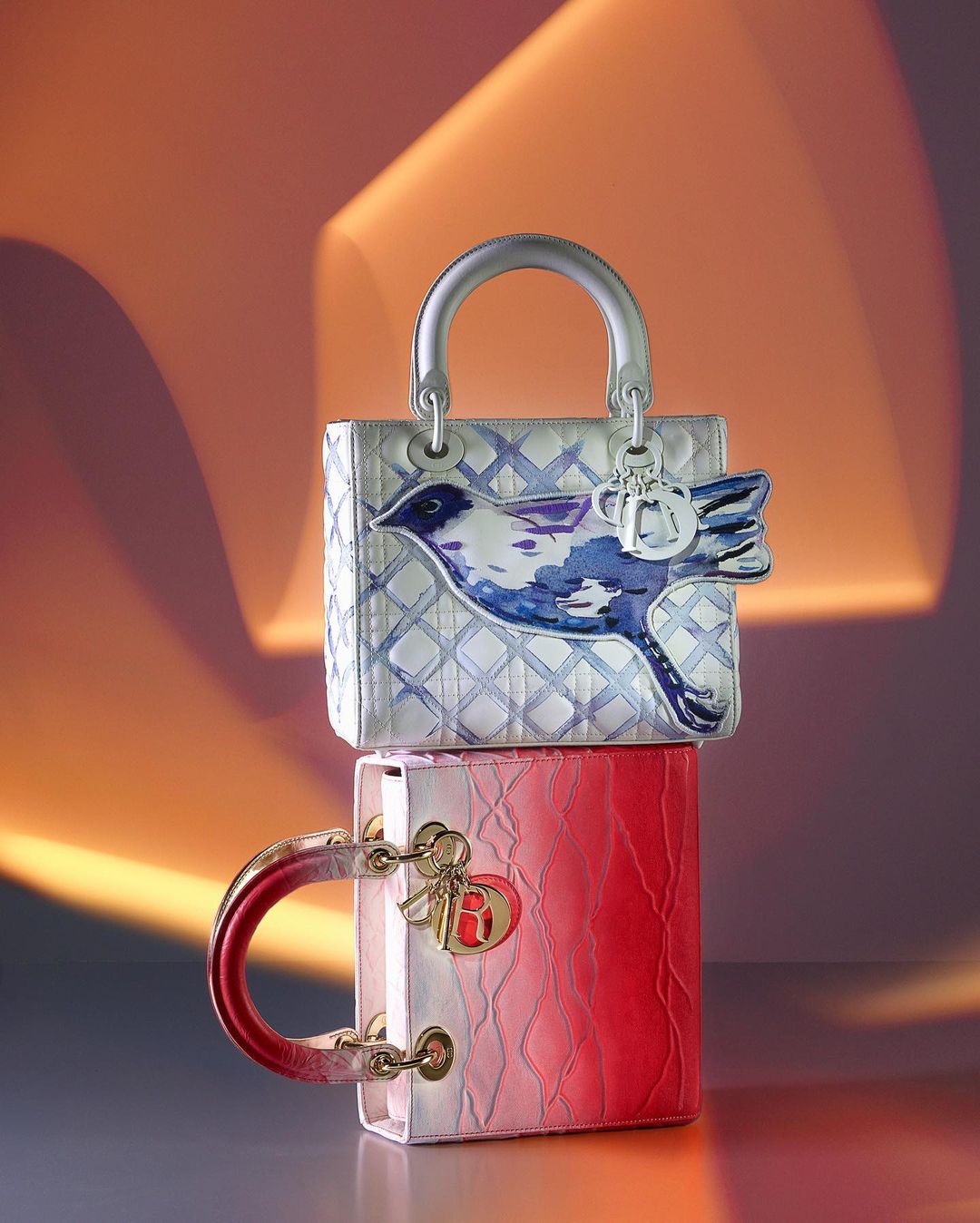 Two Lady Dior bags are stacked on top of each other, one featuring a bird design and the other an ombre red hue.
