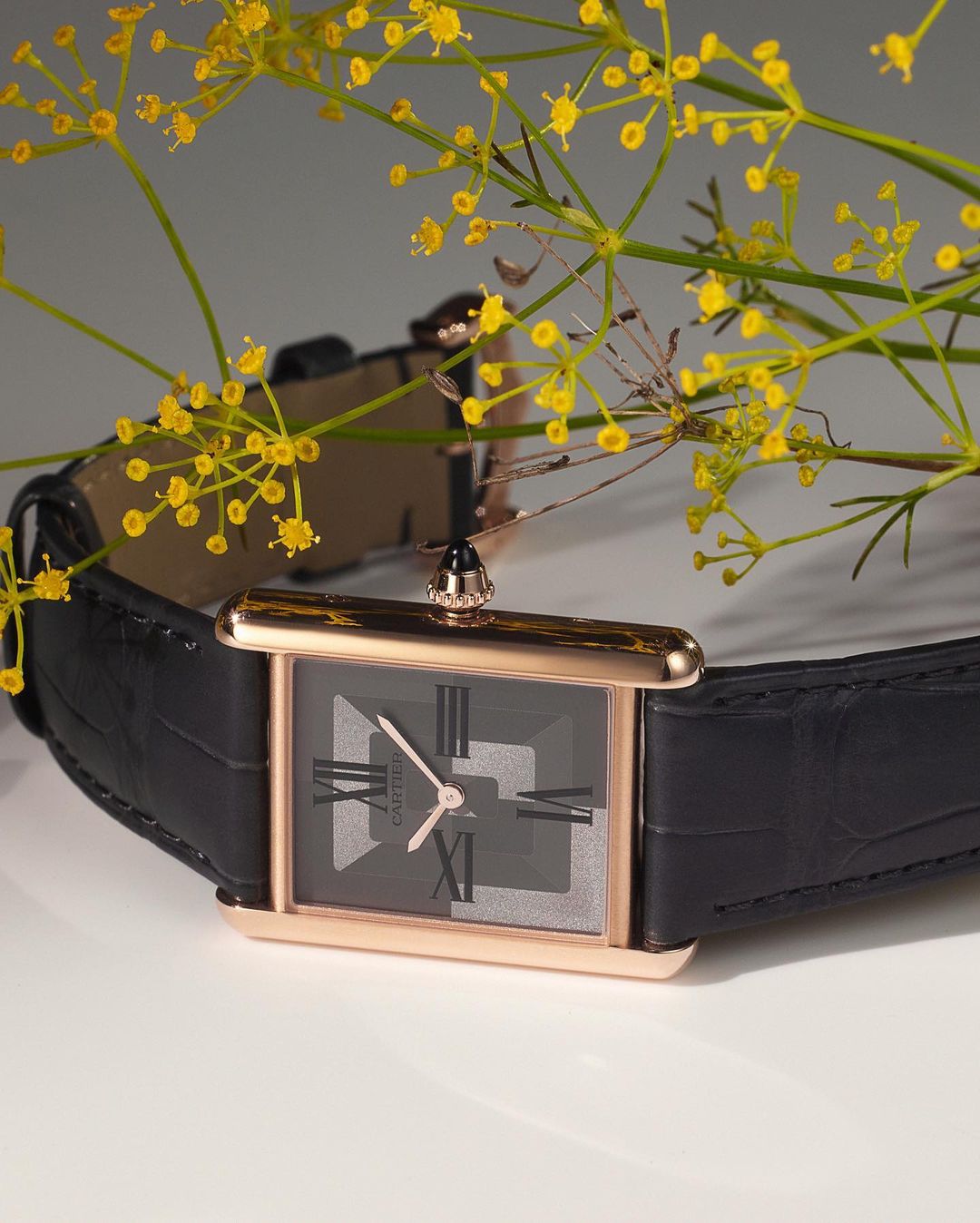 Elegant gold wristwatch with a black leather band is showcased with delicate yellow flowers, set against a neutral background. 