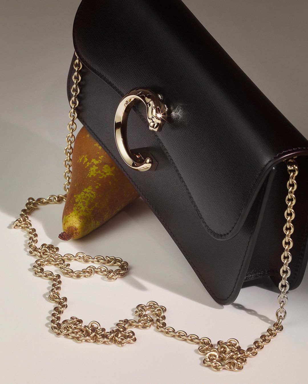 Elegant black leather clutch with a gold chain strap and panther-shaped C clasp, paired with a ripe pear on a soft cream background.