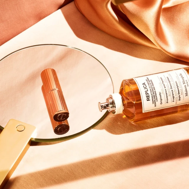 Still life of 'Replica' perfume bottle, cylindrical metal container, and round mirror on a beige backdrop with soft, diffused lighting.