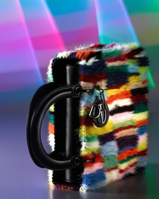 Colorful patchwork handbag with black handle and silver hardware on a gradient purple background with blurred neon light streaks.