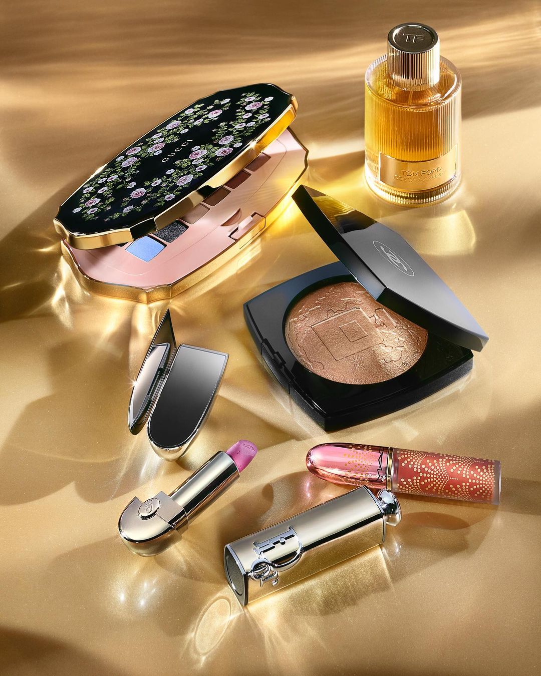 Still life shot of assorted makeup including eye shadow palettes, lipstick, and perfume on a golden surface with diffused light.