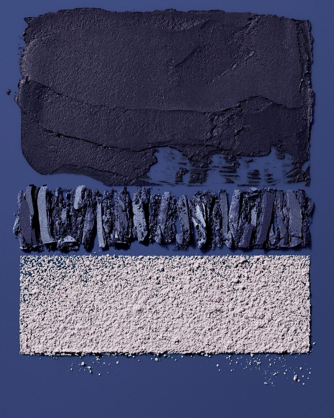 Macro studio image of dark grey eyeshadow spread, crumbled eyeshadow, and loose powder, arranged on a blue background for beauty advertising.