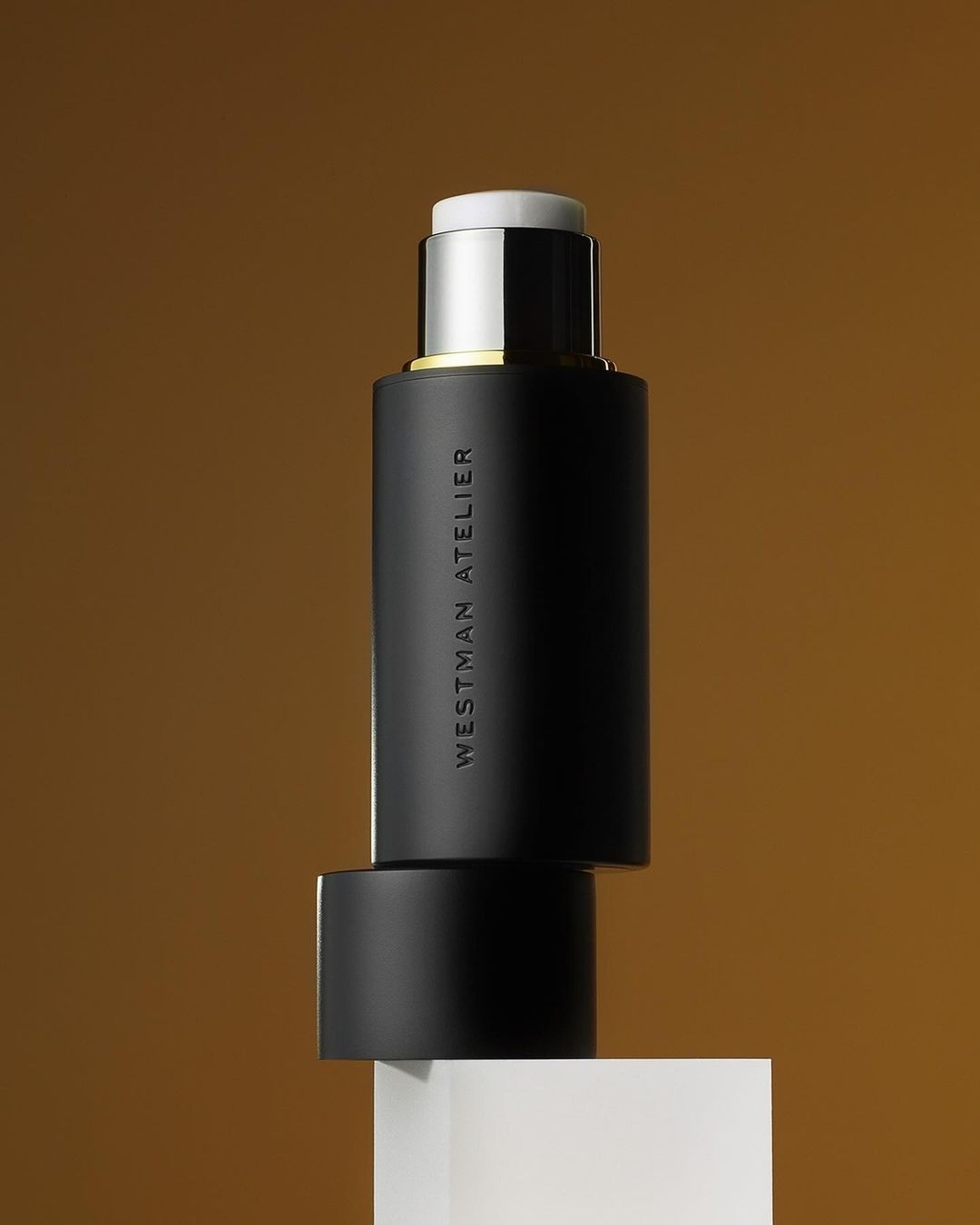 Elegant product shot of Westman Atelier makeup stick against a gradient brown backdrop, resting on a small white cube.