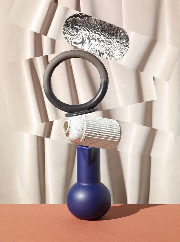 Still life arrangement featuring a blue vase, a white speckled container, a black ring, and a patterned dish against a geometric backdrop.