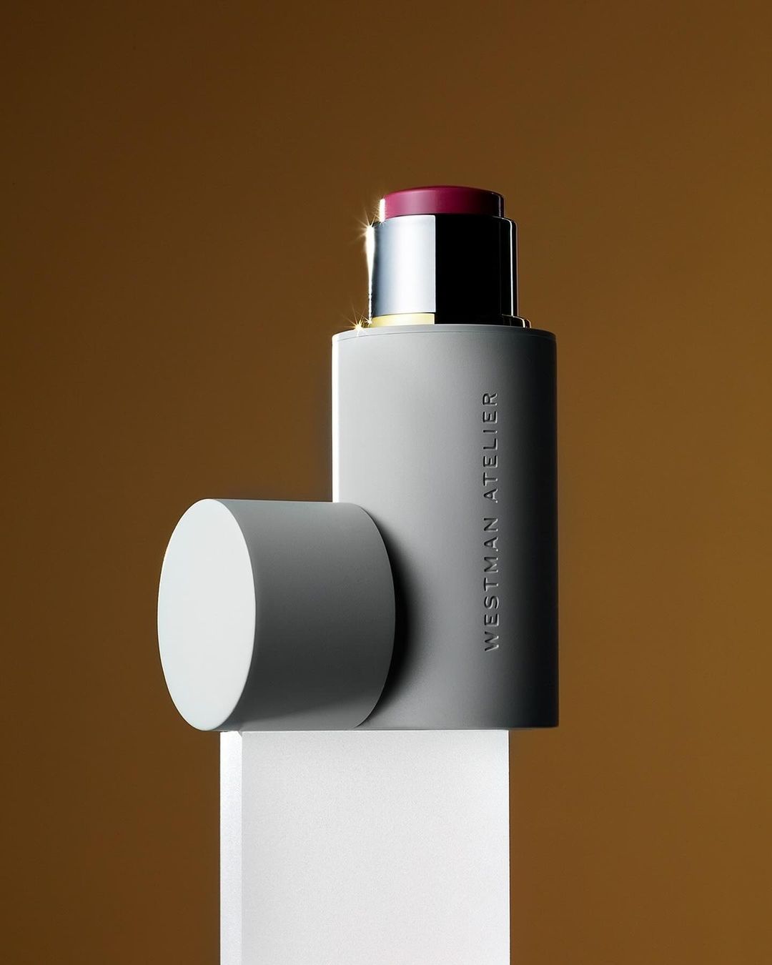 Beauty product shot: Westman Atelier makeup stick with detached lid on a white surface against a graduated brown backdrop.
