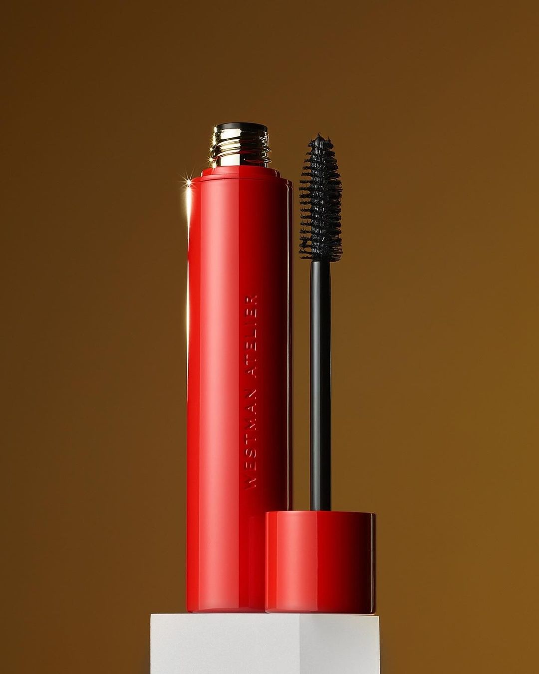 Still life studio shot of red Westman Atelier mascara tube with black wand, against a golden gradient background on a white block.