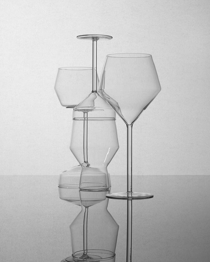 Still life featuring stacked crystal glassware; wine glasses and tumblers reflecting on a shiny surface in a grey gradient background.