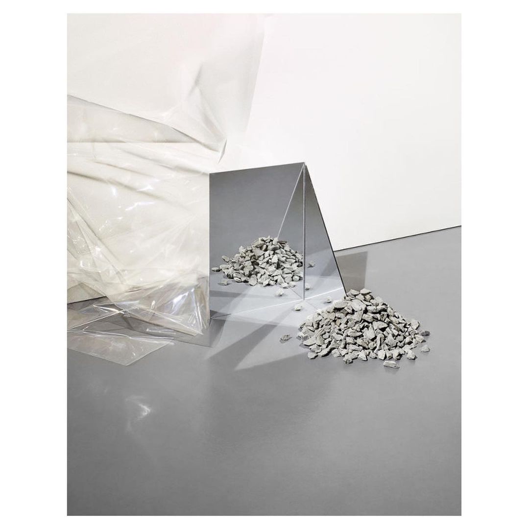 Abstract still life featuring scattered grey rocks near a reflective triangular mirror, set against white and grey surfaces.