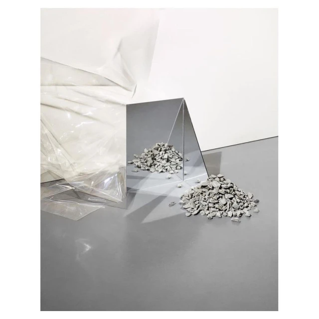 Abstract still life featuring scattered grey rocks near a reflective triangular mirror, set against white and grey surfaces.
