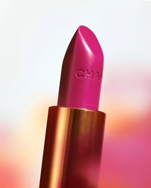 Close-up product shot of a vibrant fuchsia Chanel lipstick with a golden tube and subtle logo against a soft, blurred backdrop.