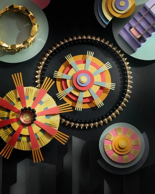 Overhead shot of colorful concentric paper circles, bracelets, and necklaces arranged on a dark background for a jewellery advertisement.