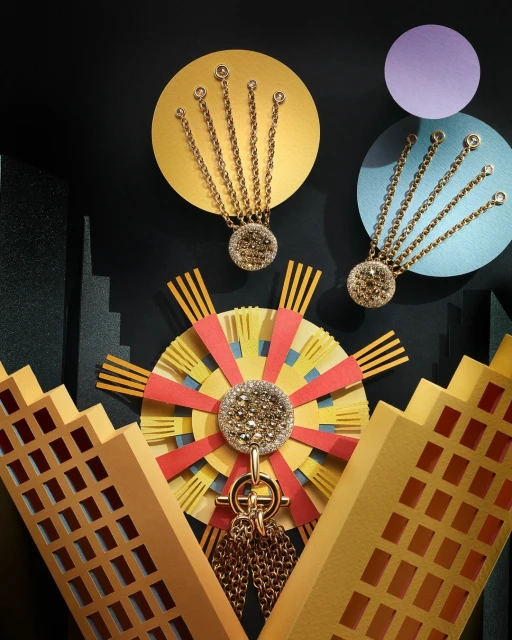 Conceptual jewelry still life featuring gold pendants with gemstone accents, against a black background with yellow, red, and blue paper shapes.