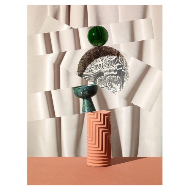 Still life of a green orb, decorative bowl, patterned disc, and textured cylinder on a terra-cotta surface against a layered paper background.
