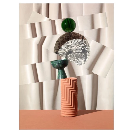 Still life of a green orb, decorative bowl, patterned disc, and textured cylinder on a terra-cotta surface against a layered paper background.