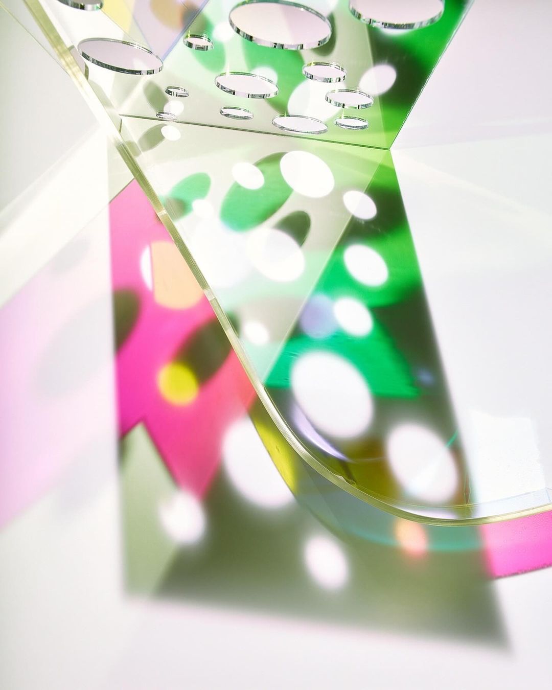 Abstract still life of curved transparent acrylic sheets casting colorful shadows with polka dots on a white surface in bright studio light.