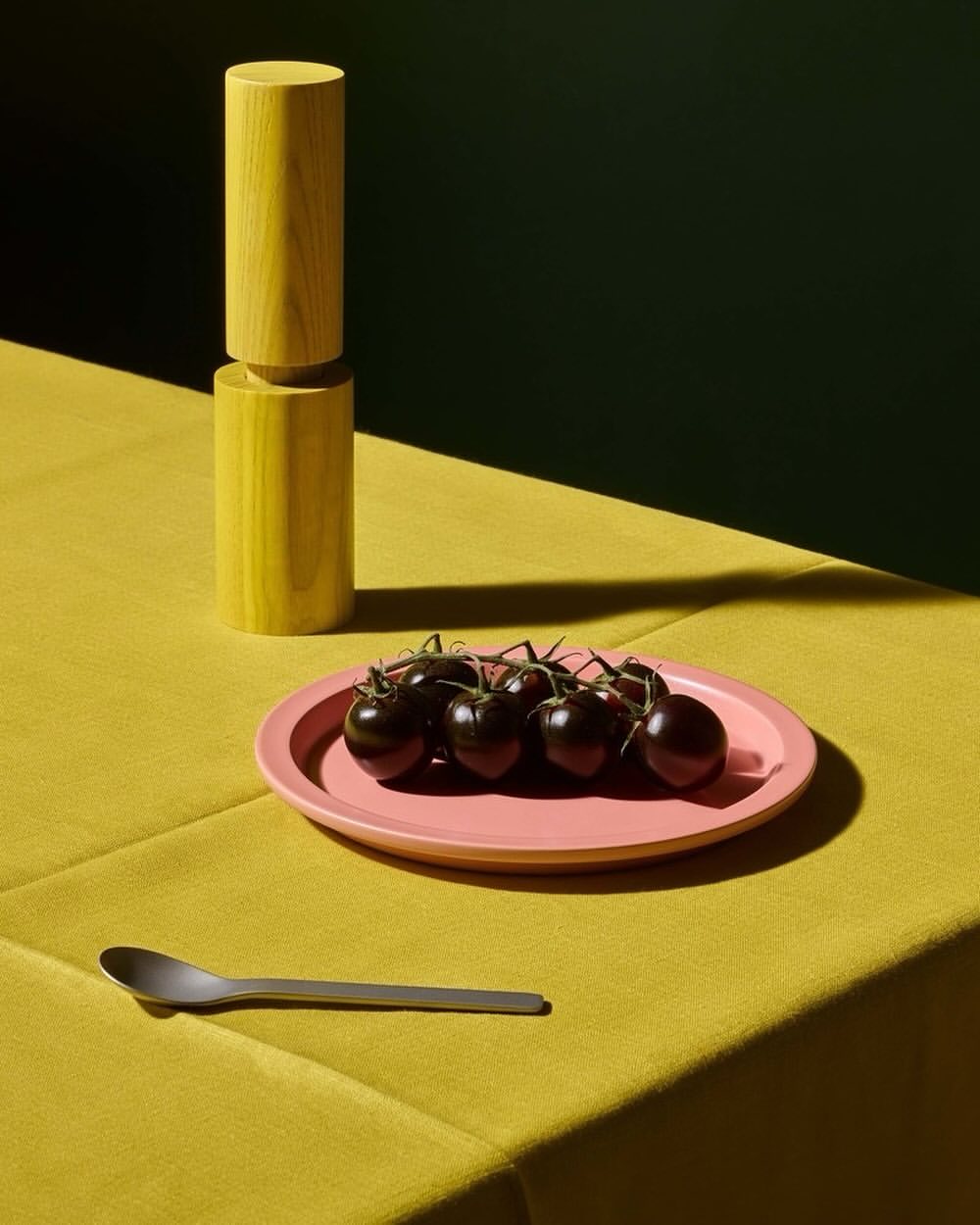 Still life shot of black tomatoes on a pink plate, yellow cylinders, a silver spoon, set against a dark green and yellow backdrop.