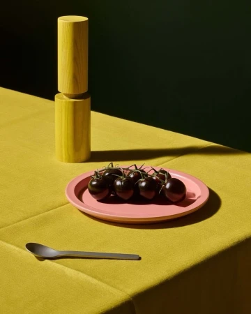 Still life shot of black tomatoes on a pink plate, yellow cylinders, a silver spoon, set against a dark green and yellow backdrop.