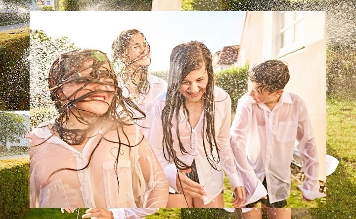 Group of four children in white shirts, soaked with water, standing outside on a sunny day with a house and green foliage in the background.
