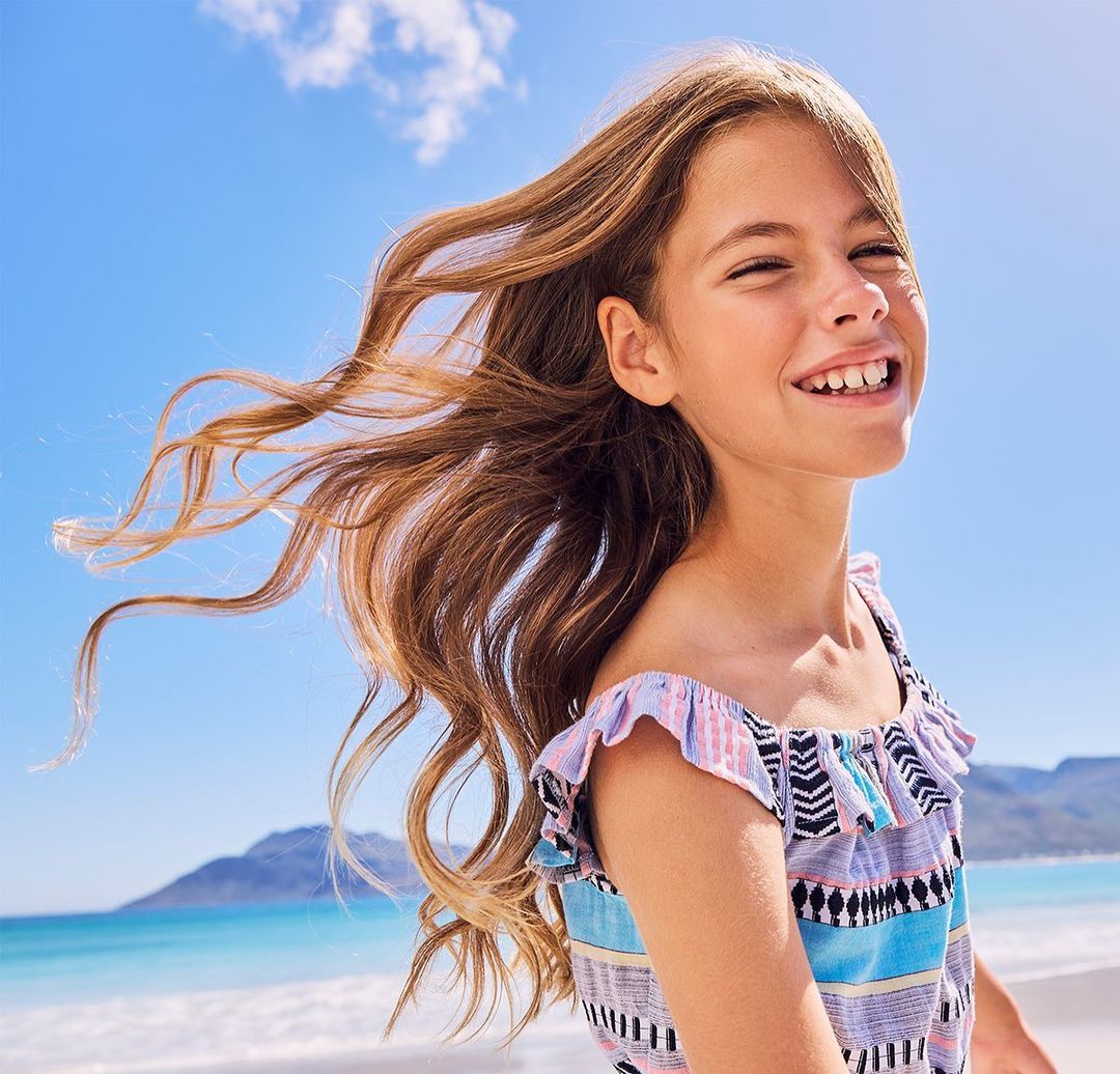 A young girl with long brown hair smiles on a sunny beach. She wears a colorful top and stands in front of the blue ocean and sky.