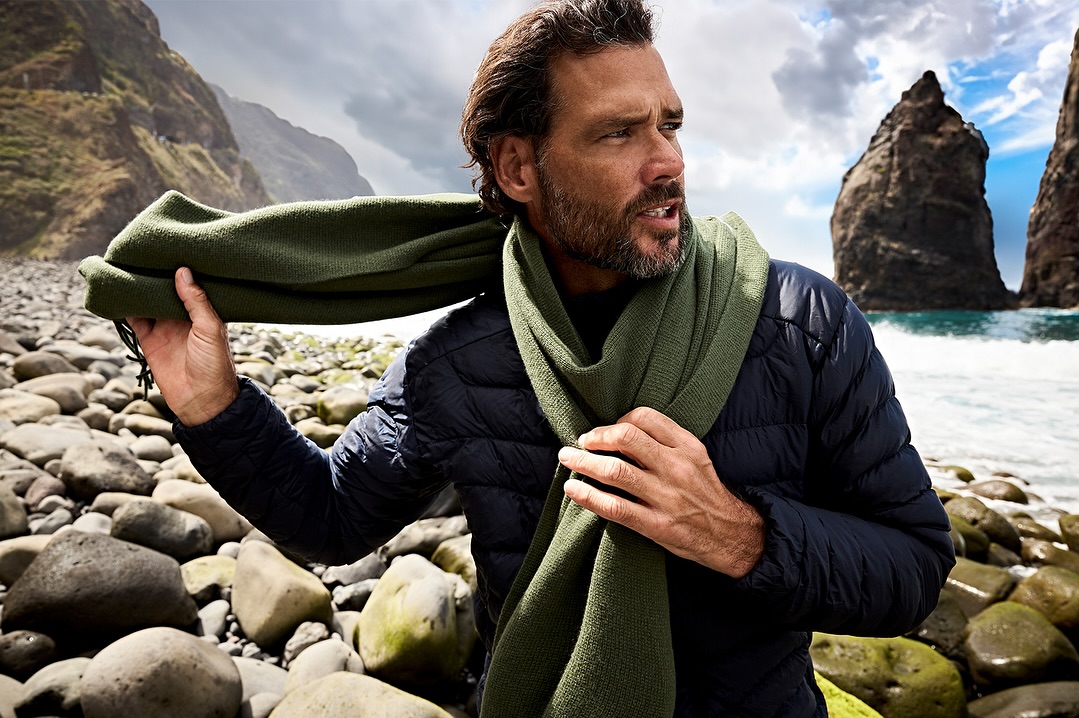 Man wearing a dark blue puffer jacket and green scarf stands on a rocky beach with dramatic rock formations in the ocean against a cloudy sky.