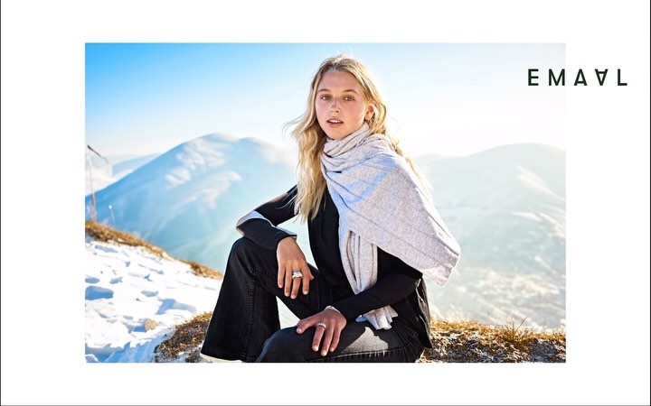 A young woman with blond hair sits on a snowy mountain slope, wearing a black shirt and a grey scarf, with mountains in the background.