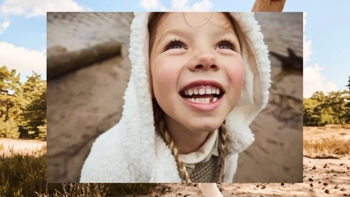 Smiling child with missing tooth wears a hooded sherpa jacket outdoors near a forest under a partly cloudy sky.