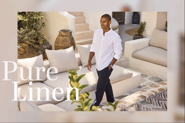 A male model walks on a stone path wearing a white shirt and dark pants in an outdoor Mediterranean setting with white buildings.