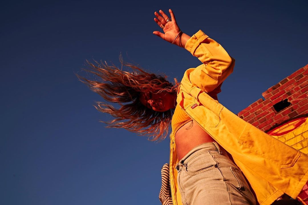 Adult female in yellow jacket raises hand to the sky, wind blows hair, low angle, blue sky background with brick structure on the right.